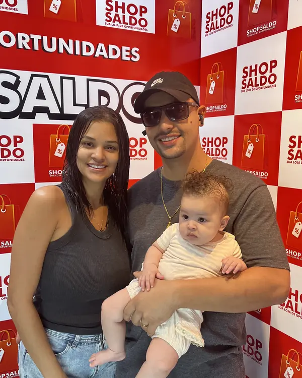 Casal com um bebê no colo posando na frente do painel vermelho da Shop Saldo