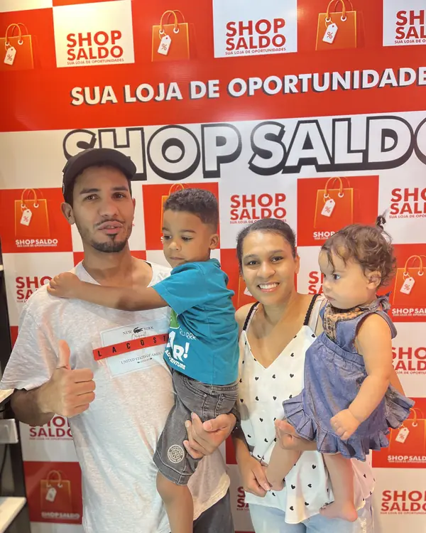 Família com duas crianças posando sorrindo na frente do painel vermelho da Shop Saldo