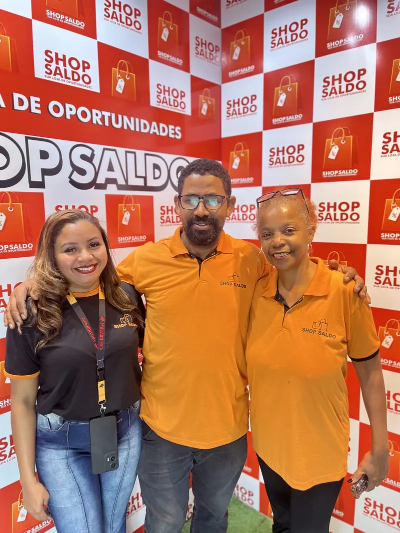 Três pessoas da equipe da Shop Saldo posando juntas na frente do painel da loja